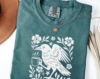May include: Teal Comfort Colors t-shirt featuring a white graphic of a crow holding a coffee cup, surrounded by floral elements. A gold necklace with a pendant is draped across the shirt. The shirt has a crew neck and short sleeves.