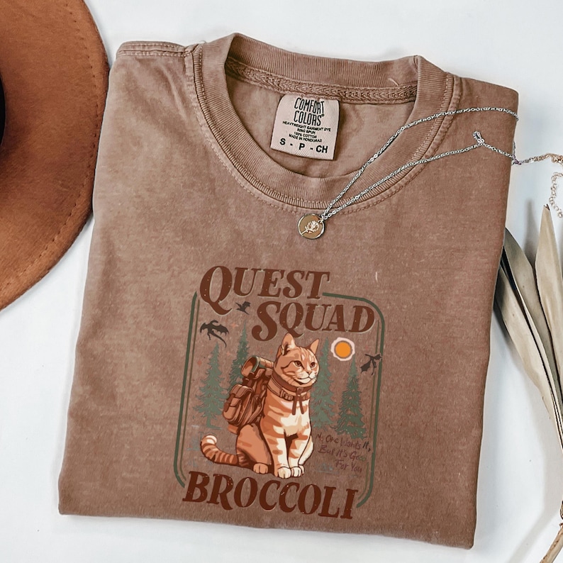 May include: A brown t-shirt with the words "QUEST SQUAD BROCCOLI" and a graphic of a cat wearing a backpack. The shirt also has a silver necklace with a small pendant. The shirt is a size small.