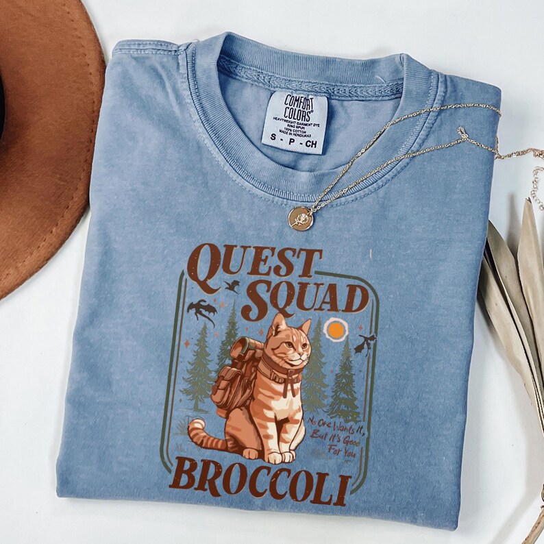 May include: A light blue Comfort Colors t-shirt with a graphic featuring a cat wearing a backpack, the words "QUEST SQUAD" and "BROCCOLI". A gold necklace is draped across the shirt.