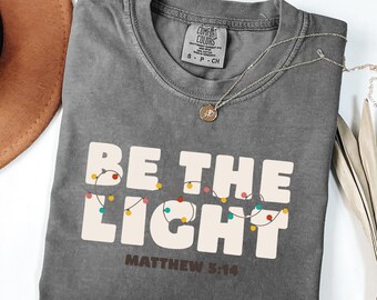 May include: A gray Comfort Colors t-shirt with the words "BE THE LIGHT" in white block letters, decorated with colorful string lights. The text "Matthew 5:14" is below the main text. A silver necklace with a pendant is on the shirt.