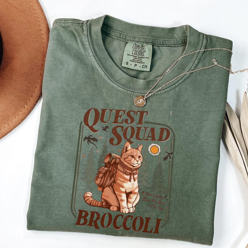 May include: Green Comfort Colors t-shirt with a graphic of a cat wearing a backpack, the words "QUEST SQUAD" and "BROCCOLI". A silver necklace with a pendant is draped across the shirt. The shirt is size S-P-CH.