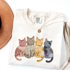 Può includere: T-shirt Comfort Colors color crema con grafica di quattro gatti stilizzati. Ogni gatto ha un motivo e un colore unici, tra cui paisley rosa, quadretti grigi, floreale giallo e patchwork blu. Sono visibili anche una collana d'oro e un cappello marrone.