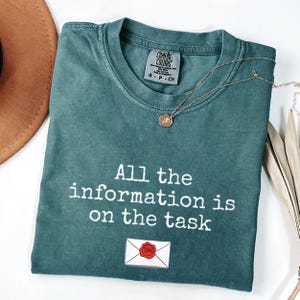 Może przedstawiać: Zielona koszulka Comfort Colors z białym napisem "All the information is on the task". Widoczny jest również złoty naszyjnik i grafika koperty. Koszulka jest złożona.