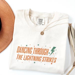 Pode incluir: Camiseta creme com o texto laranja "DANCING THROUGH THE LIGHTNING STRIKES" e um raio verde. Um colar dourado e um chapéu marrom também são visíveis. A camiseta está dobrada.