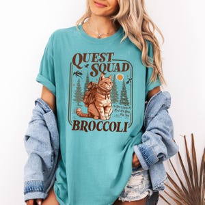 May include: Teal-colored t-shirt featuring a graphic of a cat wearing a backpack, with the words "QUEST SQUAD BROCCOLI." The design includes trees, birds, and a sun. The shirt has rolled-up sleeves.