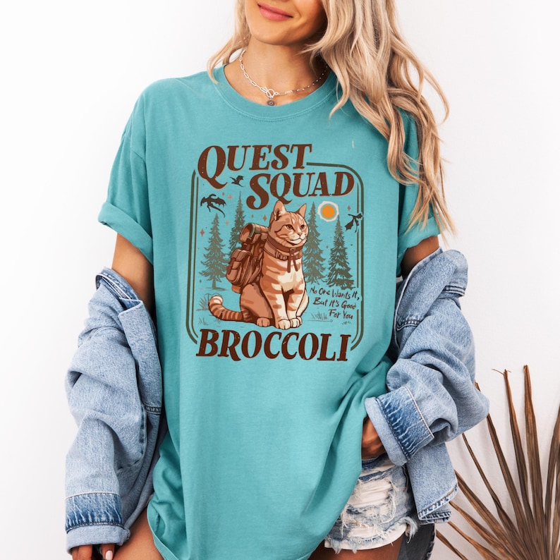 May include: Teal-colored t-shirt featuring a graphic of a cat wearing a backpack, with the words "QUEST SQUAD BROCCOLI." The design includes trees, birds, and a sun. The shirt has rolled-up sleeves.