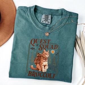 May include: Teal-colored t-shirt with a graphic of a cat wearing a backpack, the words "QUEST SQUAD BROCCOLI" are printed below the cat. A gold necklace and a brown hat are also visible.