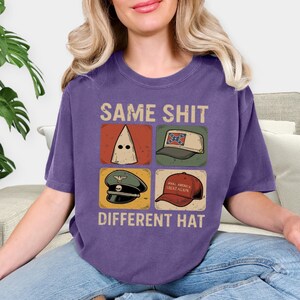 Peut inclure: T-shirt violet avec l'inscription "SAME SHIT DIFFERENT HAT" au-dessus de quatre images de chapeaux. Les chapeaux comprennent une cagoule blanche, une casquette de baseball avec un drapeau confédéré, un chapeau militaire et un chapeau rouge "Make America Great Again".