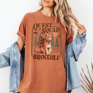 May include: Rust-colored t-shirt with a graphic featuring a cat wearing a backpack, the words "QUEST SQUAD BROCCOLI", and the phrase "No One Wants It, But It's Good For You". The design includes trees, birds, and a sun.