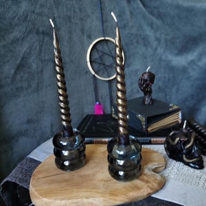 May include: Two black and gold twisted candles on glass candle holders. The candles are on a wooden cutting board.