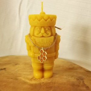 Può includere: Una candela di cera d'api gialla a forma di schiaccianoci che indossa una corona e una collana con un ciondolo a nido d'ape.