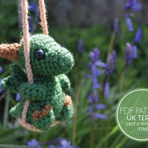 May include: A green crocheted dragon with brown accents is sitting on a swing made of rope. The dragon has one large eye visible. The dragon is in front of a background of purple flowers.