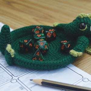 Pu&ograve; includere: Un drago di uncinetto verde che tiene un set di sei dadi verdi e neri con numeri arancioni. I dadi sono in una ciotola di uncinetto.