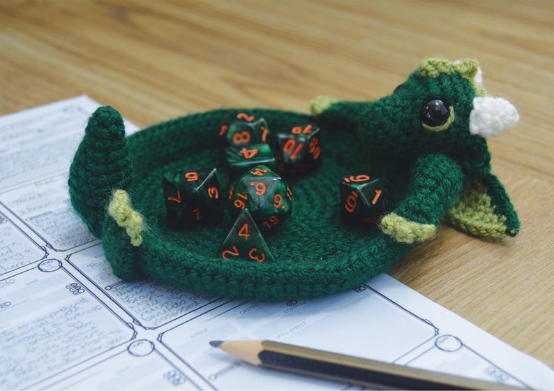Pu&ograve; includere: Un drago di uncinetto verde che tiene un set di sei dadi verdi e neri con numeri arancioni. I dadi sono in una ciotola di uncinetto.