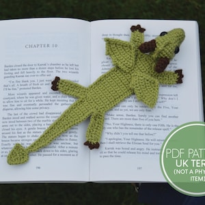 以下が含まれることがあります： 茶色の目と羽根を持つ緑色の編みぐるみドラゴンブックマーク。ブックマークは、「第10章」というテキストが見える開いた本の上に置かれています。ブックマークは、編み物のPDFパターンです。