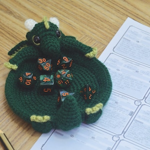 Pu&ograve; includere: Sacchetto da gioco per dadi in crochet verde a forma di drago con bordo giallo e diversi dadi neri e arancioni all'interno.