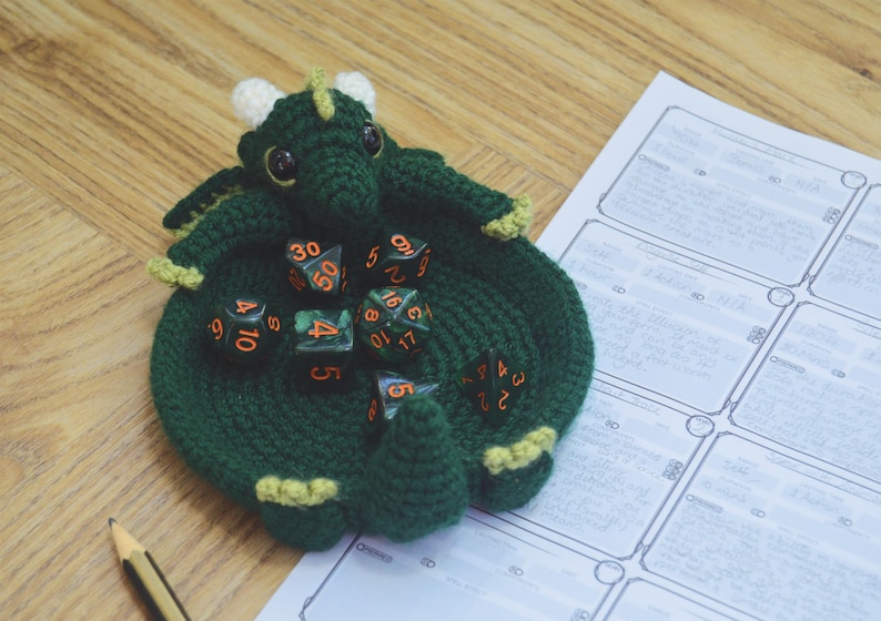 Pu&ograve; includere: Sacchetto da gioco per dadi in crochet verde a forma di drago con bordo giallo e diversi dadi neri e arancioni all'interno.