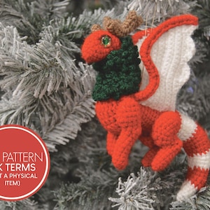 May include: A handmade crochet dragon Christmas ornament. The dragon is red with white and green accents, and brown antlers. The wings are white. The ornament is hanging on a frosted Christmas tree. The text in the image says "PDF PATTERN UK TERMS (NOT A PHYSICAL ITEM)".