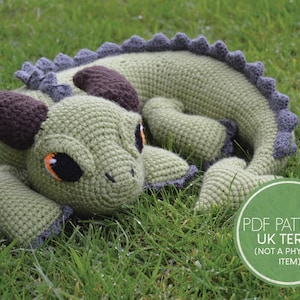 May include: A green crocheted dragon with brown horns and orange eyes. The dragon is lying on its side on a bed of green grass. The text "PDF PATTERN UK TERMS (NOT A PHYSICAL ITEM)" is visible in the lower right corner of the image.