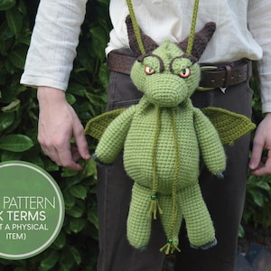 May include: A green crocheted dragon bag with brown horns, wings, and orange eyes. The dragon has a drawstring closure and a long strap. The bag is being held by a person. A green circle with white text that reads "PDF PATTERN UK TERMS (NOT A PHYSICAL ITEM)" is in the lower left corner.