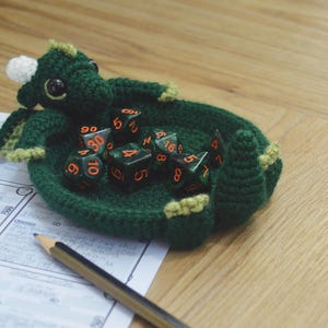 Pu&ograve; includere: Vassoio da gioco per dadi a forma di drago in crochet verde con un corno bianco e dettagli gialli. Il vassoio &egrave; pieno di dadi verdi e arancioni.