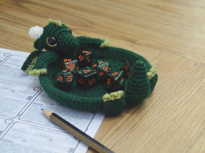 Pu&ograve; includere: Vassoio da gioco per dadi a forma di drago in crochet verde con un corno bianco e dettagli gialli. Il vassoio &egrave; pieno di dadi verdi e arancioni.