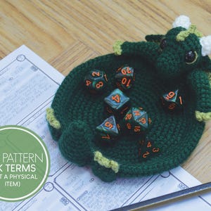 Könnte beinhalten: Grüner gehäkelter Drachen-Würfelhalter mit einem weißen gehäkelten Horn und grünen Würfeln mit orangenen Zahlen. Der Würfelhalter liegt auf einem weißen Blatt Papier mit einem grünen Kreis, auf dem "PDF PATTERN UK TERMS (NOT A PHYSICAL ITEM)" steht.