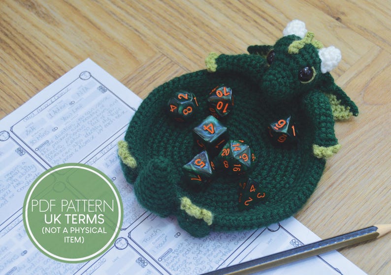 Pu&ograve; includere: Porta dadi di drago in crochet verde con un corno in crochet bianco e dadi verdi con numeri arancioni. Il porta dadi &egrave; su un foglio di carta bianco con un cerchio verde che dice "PDF PATTERN UK TERMS (NOT A PHYSICAL ITEM)".