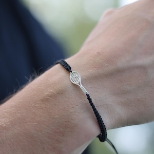 May include: A black woven bracelet featuring a silver tennis racket charm. The charm has intricate detailing, and the bracelet is secured with a sliding knot closure. The bracelet is worn on a wrist.