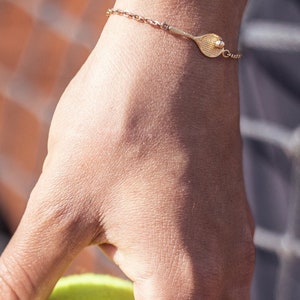 Peut inclure: Un bracelet en or avec un pendentif en forme de raquette de tennis et un petit pendentif en forme de balle de tennis. Le bracelet est porté au poignet.