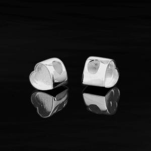 Könnte beinhalten: Zwei silberne herzförmige Perlenabstandshalter mit einem Loch in der Mitte. Die Abstandshalter befinden sich auf einer schwarzen Oberfläche.