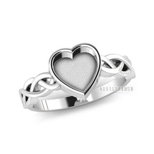 May include: A silver ring with a heart-shaped setting. The heart is framed by a smooth, polished border. The band features an interwoven Celtic knot design. The ring is set against a white background.