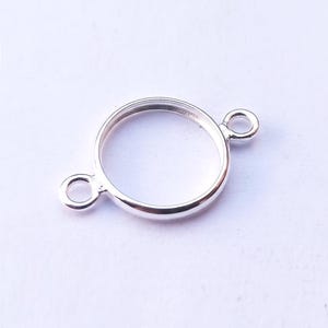 Puede incluir: Un brillante componente de joyería circular plateado con dos pequeños bucles. El componente tiene forma de anillo con una superficie lisa y pulida. Los bucles están unidos al anillo, uno a cada lado, para la conexión.