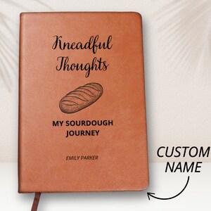 Puede incluir: Un diario de cuero marrón con el texto "Kneadful Thoughts My Sourdough Journey" y un dibujo de una hogaza de pan. El texto "Emily Parker" está debajo del dibujo. El texto "CUSTOM NAME" está en el lado derecho de la imagen.