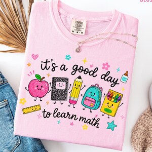 Può includere: Una t-shirt rosa chiaro con una stampa grafica con il testo "it's a good day to learn math" circondata da illustrazioni colorate di una mela sorridente, un quaderno, una matita, uno zaino, una scatola di pastelli, un righello e colla.