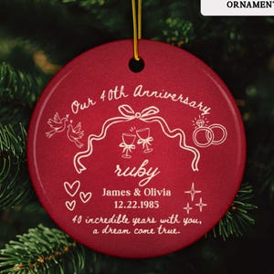 Puede incluir: Adorno redondo de cerámica roja con una cuerda dorada para colgar. Texto e ilustraciones en blanco: "Our 40th Anniversary", "ruby", nombres, fecha y mensaje. Un recuerdo personalizado.