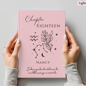 May include: A light pink journal held by two hands. The cover features the words "Chapter EIGHTEEN" and "NANCY" in black script, along with botanical and celestial illustrations. The journal's text reads "Embrace your freedom & become the incredible person you're meant to be."