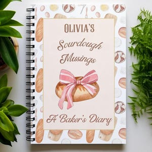 May include: A white spiral bound journal with a pink and brown illustration of a loaf of bread with a pink ribbon. The cover has a white background with a repeating pattern of bread, flour, and eggs. The text "Olivia's Sourdough Musings A Baker's Diary" is printed on the cover.