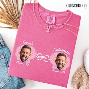 May include: A pink crewneck t-shirt featuring two printed graphics of a person's face inside a seashell, connected by a white bow illustration. The text reads Emma's Miami, FL and Bachelorette August 2026. A gold chain necklace rests on the collar.