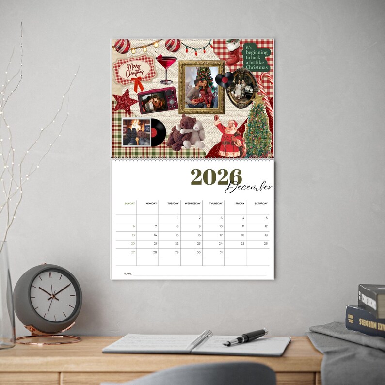 May include: A 2026 December wall calendar with a festive Christmas collage at the top. The collage includes framed photos, a teddy bear, a Santa figurine, and holiday decorations. The calendar is displayed on a wooden desk.