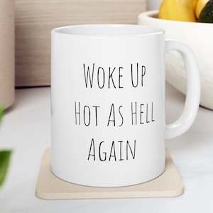 May include: White ceramic coffee mug with black text that reads "Woke Up Hot As Hell Again".