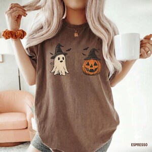 May include: A brown short-sleeve t-shirt featuring a graphic print of a ghost and a jack-o'-lantern, both wearing black witch hats. The design includes small black bats and stars surrounding the figures. The shirt has a crew neckline.