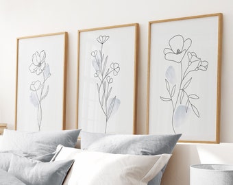 May include: Three framed line drawings of flowers in shades of gray and white on a white wall above a gray and white bed with pillows and a throw blanket.