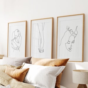 May include: Three framed black and white line drawings of couples in various poses. The drawings are hung on a white wall above a bed with brown linen bedding.