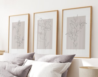 May include: Three framed line drawings of flowers and leaves in black and white on a light pink background. The frames are natural wood and the drawings are minimalist in style. The frames are hanging on a white wall above a bed with gray and white bedding.