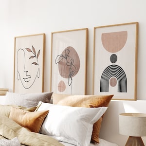 May include: Three minimalist art prints with abstract line drawings and muted colors. The prints are framed in natural wood and hung on a white wall above a bed with brown and white bedding.