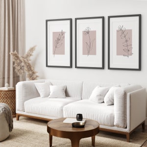 May include: Three black framed prints with minimalist line drawings of flowers and leaves on a blush pink background. The prints are hanging on a white wall above a white sofa.