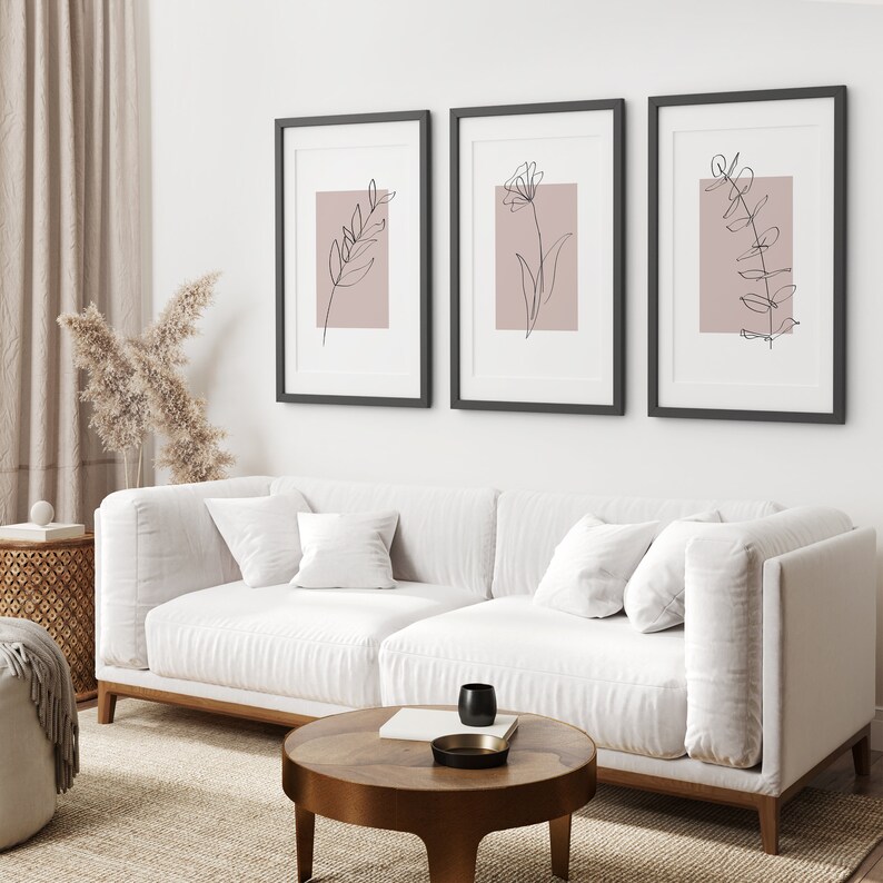 May include: Three black framed prints with minimalist line drawings of flowers and leaves on a blush pink background. The prints are hanging on a white wall above a white sofa.