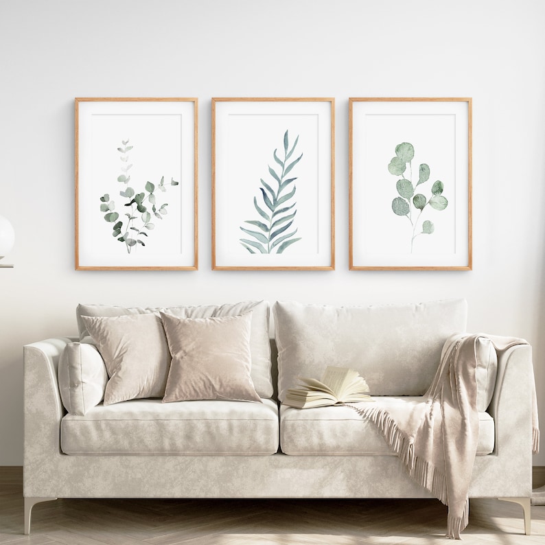 May include: Three framed watercolor prints of green eucalyptus leaves on a white wall above a light gray couch with two beige pillows and a beige throw blanket.
