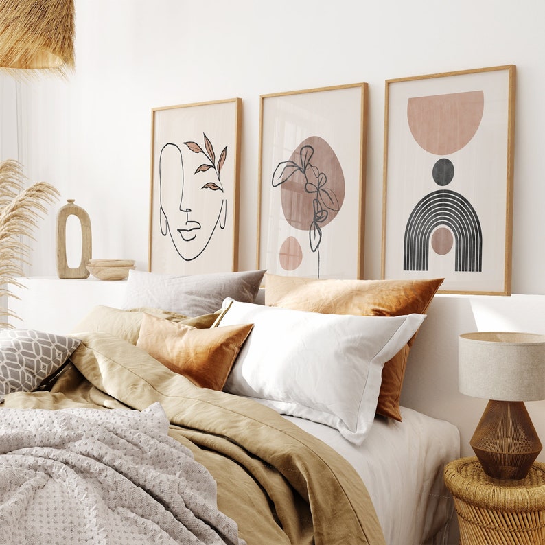 May include: Three minimalist art prints with abstract line drawings and muted colors. The prints are framed in natural wood and hung on a white wall above a bed with brown and white bedding.